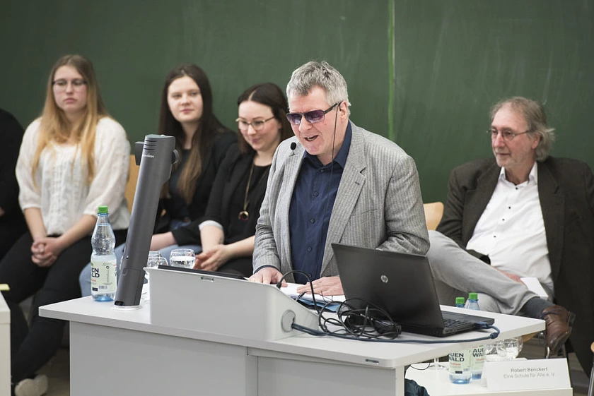Das Foto wurde im März 2019 während der Veranstaltung "Schulische Inklusion in Bremen – Bilanz und Perspektiven" gemacht. Es zeigt Joachim Steinbrück während seines Vortrag. Er steht an einem Rednerpult. Hinter ihm sitzen drei Schülerinnen und ein Lehrer.