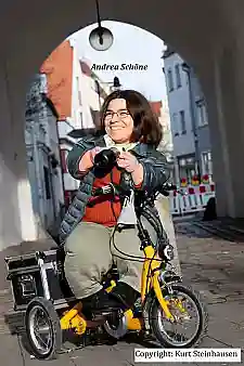 Portrait von Andrea Schöne im Sonnenschein auf ihrem Fahrrad mit Kamera unter einem Torbogen