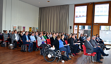 Das Foto zeigt die Teilnehmerinnen und Teilnehmer der Veranstaltung. Im Vordergrund ist eine Person im Rollstuhl zu sehen. Der Konferenzraum ist durch seine großen Fenster sehr hell.