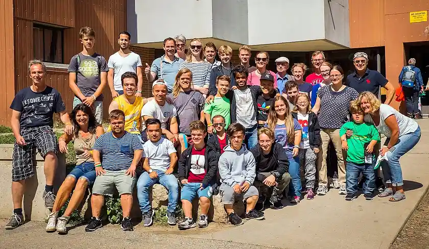 Auf dem Foto sind die Teilnehmerinnen und Teilnehmer an den World Dwarf Games sowie Angehörige abgebildet. Insgesamt zeigt das Gruppenfoto ca. 40 Personen in sommerlicher Freizeitbekleidung.