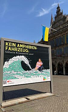 Plakat auf dem Marktplatz - Eine junge Frau im Rollstuhl sitzt vor schwarzem Hintergrund im Wasser, eine riesige Welle rollt auf sie zu. Die Frau ist real, die Welle gezeichnet. Über dem Bild steht der Text Kein Amphibienfahrzeug. Inklusive Krisenpolitik jetzt! Darunter das Logos des Landesbehindertenbeauftragten und ein QR-Code mit dem Text Scan mich, der zu dieser Webseite führt.