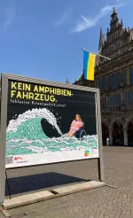 Plakat auf dem Marktplatz - Eine junge Frau im Rollstuhl sitzt vor schwarzem Hintergrund im Wasser, eine riesige Welle rollt auf sie zu. Die Frau ist real, die Welle gezeichnet. Über dem Bild steht der Text Kein Amphibienfahrzeug. Inklusive Krisenpolitik jetzt! Darunter das Logos des Landesbehindertenbeauftragten und ein QR-Code mit dem Text Scan mich, der zu dieser Webseite führt.