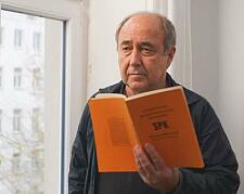Filmszene: Ein traurig dreinblickender Mann mit Halbglatze hält ein oranges Buch mit dem Titel SPK