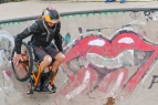 Ein Rollstuhlfahrer mit Pflaster auf den Knien skatet in der Halfpipe vor einem Graffititi der Rolling-Stones-Zunge 