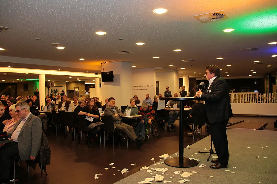 Dr. Hess-Grunewald w&auml;hrend seines Gru&szlig;wortes. Er steht auf der B&uuml;hne vor einem Stehtisch.