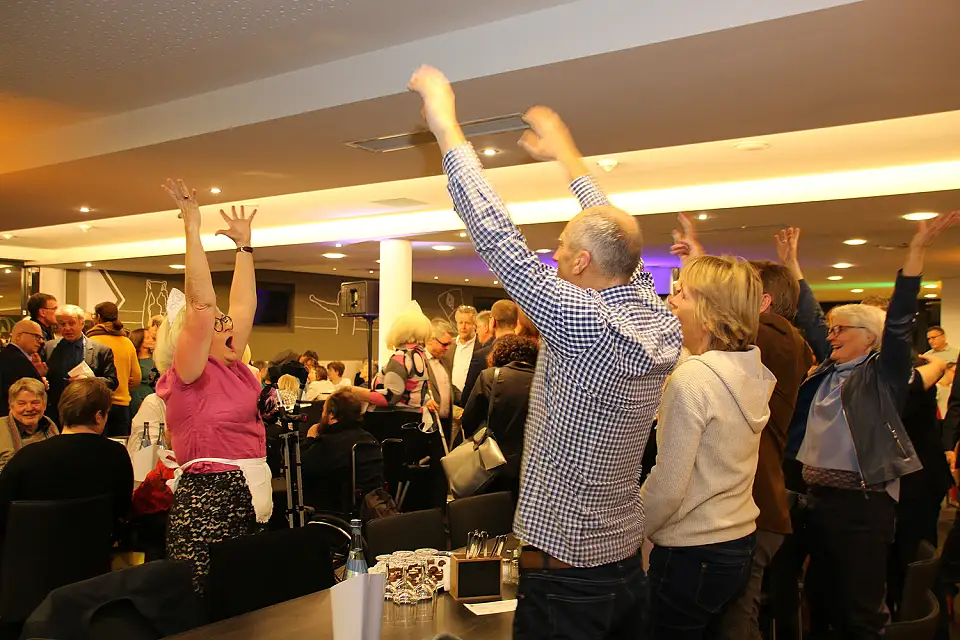 Eine "S&uuml;&szlig;e Frau" annimiert Personen an einem Tisch zu einer La-Ola-Welle